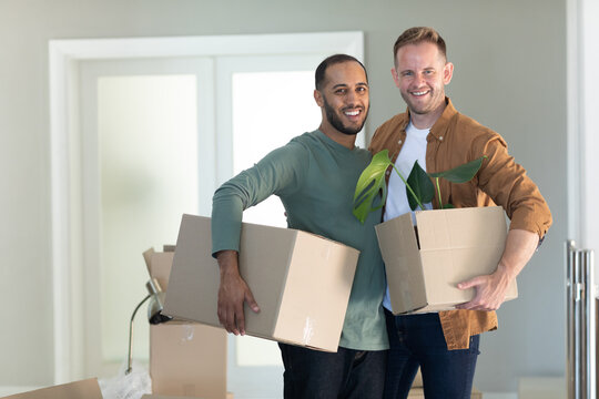 Multi ethnic gay male couple holding boxes looking at camera and smiling at home