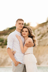 A beautiful young couple in love, a man and a woman embrace, kiss near a blue lake and sand at sunset. Vacation at the sea on the beach, sunset, morning, honeymoon, tropics, ocean, girl, relationship.