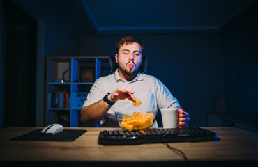 Hungry bearded man eating chips from a plate at night at the computer at home in a room with blue light. Freelancer eats chips during night work.