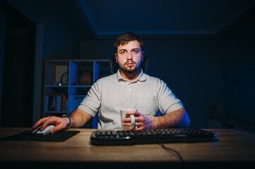 Handsome man freelancer with a beard sits at night at his desk at the computer and works with a cup of drink in his hand, looks at the computer screen and works.