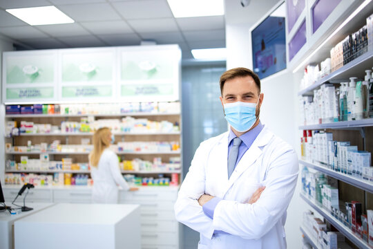 Portrait of pharmacist wearing face mask and white coat standing in pharmacy store during corona virus pandemic.