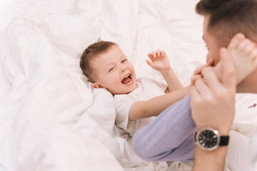 Young dad playing and laughing with his baby smiling son at home in bed in the bedroom, happy family