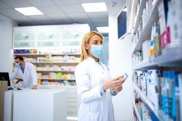 Vrouwelijke apotheker met gezichtsmasker en witte jas die medicijnen vasthoudt in de apotheek tijdens de pandemie van het coronavirus.