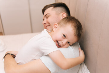 Obraz na płótnie Canvas Young dad playing and laughing with his baby smiling son at home in bed in the bedroom, happy family