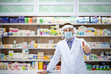 Portrait of pharmacist wearing face shield and white coat standing in pharmacy store during corona virus pandemic.