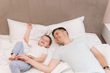 Young dad playing and laughing with his baby smiling son at home in bed in the bedroom, happy family