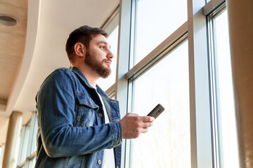 Fototapeta premium Man use smartphone while standing near window in beautiful modern business center. Young man in denim jacket near panoramic window with phone. Man texting message via smartphone.