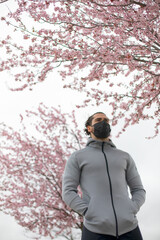 Muscular and handsome young man with sportswear under a cherry blossom tree. The dark haired athlete is wearing a mask.