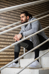 Young bearded athlete with dark hair having a break from working out on a cold day and sitting on metalic stairs