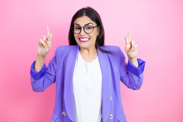 Young business woman wearing purple jacket over pink background gesturing finger crossed smiling with hope and looking side