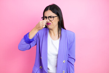 Young business woman wearing purple jacket over pink background smelling something stinky and disgusting, intolerable smell, holding breath with fingers on nose