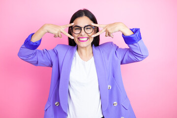 Young business woman wearing purple jacket over pink background Doing peace symbol with fingers over face, smiling cheerful showing victory
