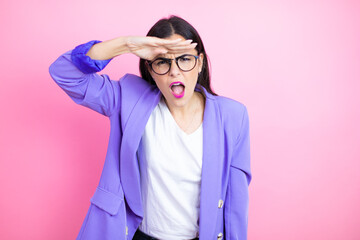 Young business woman wearing purple jacket over pink background very happy and smiling looking far away with hand over head. searching concept.