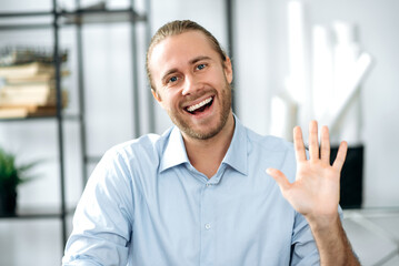 Webcam portrait of a happy caucasian freelancer guy during video conference, he looks at the camera waving hand and smiling, greeting his friends, family or colleagues