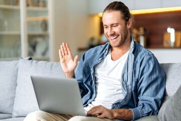 Joyful male freelancer using laptop, have a video call, communicate with colleagues or friends by video conference, waving hand and smiling. Handsome caucasian man is sitting on a sofa, working from