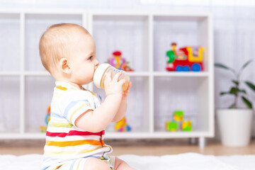 Cute caucasian baby boy drinking from bottle fruit tea with camomile. Sitting on white carpet in striped bodysuit. Different children's toys in the background. Side view