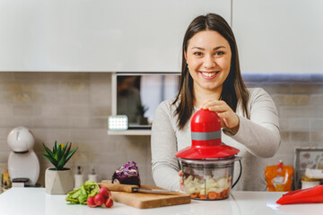 Front view portrait of adult happy caucasian woman smile while using blender to prepare vegetables...