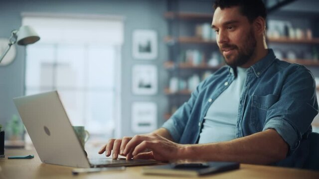 Handsome Caucasian Man Working on Laptop Computer while Sitting on a Sofa Couch in Stylish Cozy Living Room. Freelancer Working From Home. Browsing Internet, Using Social Networks, Having Fun.