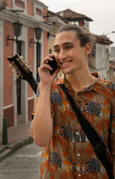 Hispanic Young Man In The Center Of Bogota Colombia, La Candelaria, A Place Of Colonial Houses Perfect For A Tourist Destination, Talking On A Cell Phone, Holding A Guitar