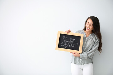 Young female teacher holding chalkboard with words DO YOU SPEAK ENGLISH? on light background. Space...