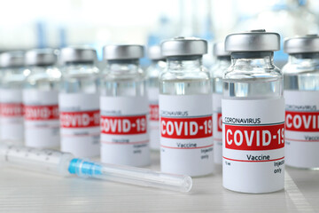 Glass vials with COVID-19 vaccine and syringe on white wooden table