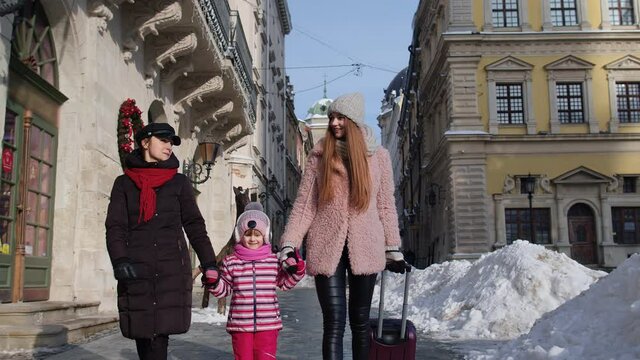 Two young smiling women travelers with adoption child girl walking with suitcase, looking at famous sights of old city streets. Lesbian couple talking, smiling outdoor. Winter holidays vacation trip