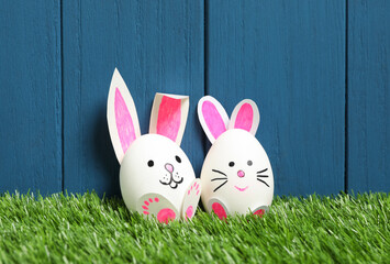 Bright Easter eggs as cute bunnies on green grass against blue wooden background