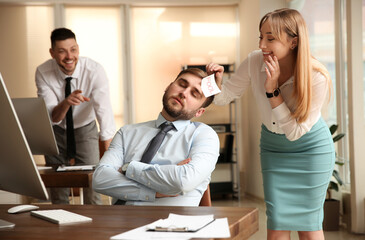 Young woman sticking note with word Fool to colleague's face in office. Funny joke