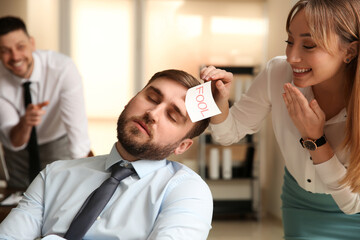 Young woman sticking note with word Fool to colleague's face in office. Funny joke
