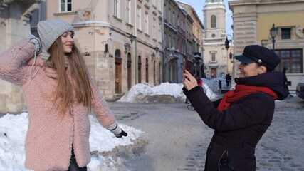 Fototapeta premium Two young smiling women tourists bloggers taking photos portrait on mobile phone while standing on city street. Happy lesbian LGBT couple during winter holidays traveling vacation making online vlog