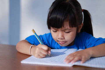 A cute little Asian girl is writing a book with a pencil on the table.