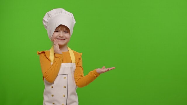 Funny preschooler child girl dressed cook chef baker in apron and hat pointing at right on blank space on chroma key background. Place for your advertisement logo. Cooking school, children education
