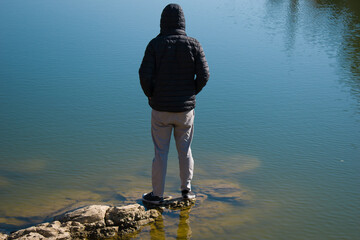 boy standing on shore of lake on rocks watching beauty of lake wearing black hood