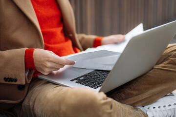 Close up of man wear wear red sweater and beige coat holding and reading documents, remote work with laptop, sitting outdoors. Young entrepreneur male distance working on computer. 