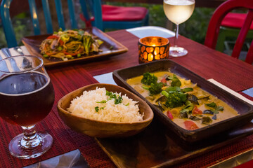 vegan cuisine with drinks on a table set for two. These healthy options are Thai Green Curry with white rice and Pad Thai. Glasses of white wine and beer are also on the candlelit table