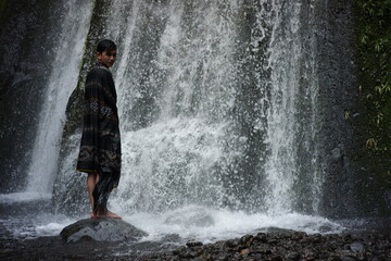 A portrait of an Asian man wearing a Lombok-Indonesian songket cloth enjoying the freshness of a waterfall in the middle of the forest. a traveler relaxing and bathing under a swift waterfall
