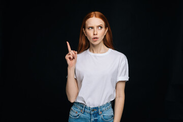 Red-haired young woman wearing T-shirt and denim having good idea with her finger pointing up isolated black background. Pretty redhead lady model emotionally showing facial expressions.