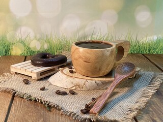 An oak table with visible grains is standing on a wood cup with black coffee. On a jute napkin, coffee beans are scattered and a bitten gingerbread heart lies on a birch pad 
