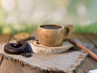 An oak table with visible grains is standing on a wood cup with black coffee. On a jute napkin, coffee beans are scattered and a bitten gingerbread heart lies on a birch pad 