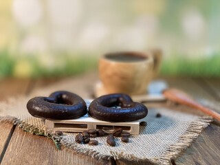 An oak table with visible grains is standing on a wood cup with black coffee. On a jute napkin, coffee beans are scattered and a gingerbread bitten heart lies on the pallet 