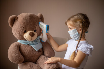 A little girl with pigtails in a mask measures the temperature of a large toy bear in a scarf, treats, plays a child's doctor. Healthcare concept, vaccination of children, pediatrician. Selective focu