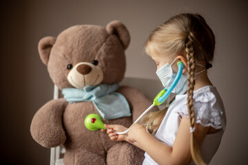 Little girl with pigtails in a mask Listens with a phonendoscope to a big toy bear in a scarf, treats, plays a child's doctor. Healthcare concept, vaccination of children, pediatrician. Selective focu