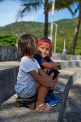 Smiling little boy with baseball cap looks at camera and sits with his sister on skateboard outside. Summertime weekend in Thailand.