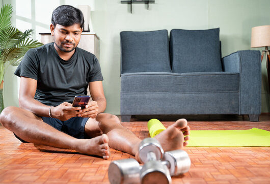 Wide Angle Shot Of Young Man Busy Using Mobile Phone During Work Out At Home - Millennial Checking Online Exercise Tutorials For Workout