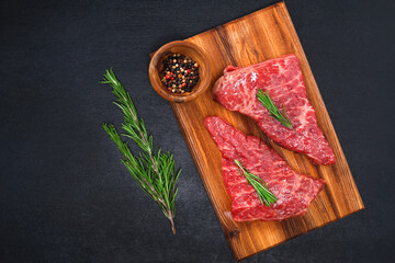 Raw steak on a cutting board with rosemary and spices, dark black background, top view. Fresh grilled meat. Grilled beef steak.