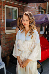 bride in a white silk robe stands in the yard of her house