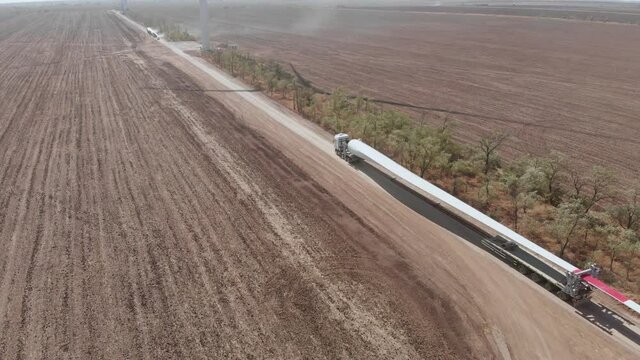 Powerful truck transports long blade for wind turbine along rural road past empty fields on sunny day under blue sky aerial view