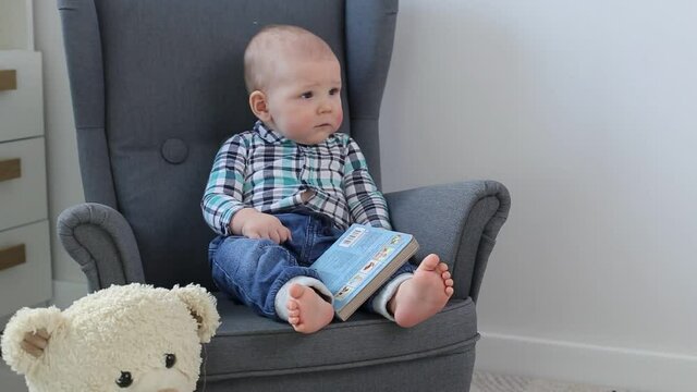A cute little baby in a shirt sits in a child's chair with a picture book.
