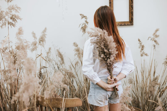 red haired girl closes her face with bouquet wearing a white shirt and denim shorts in a space decorated with dry grass and flowers in a retro style