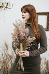 young woman plus size model with handmade candles from wax in a green dress stands in a space decorated with dry grass and flowers in a retro style